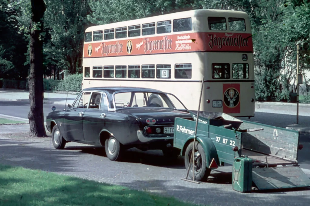 Ein altes Foto aus den frühen Jahren. Zu sehen ist ein Auto samt Anhänger samt Aufschrift mit der "R. Fehmer", im Hintergrund fährt ein Bus vorbei.