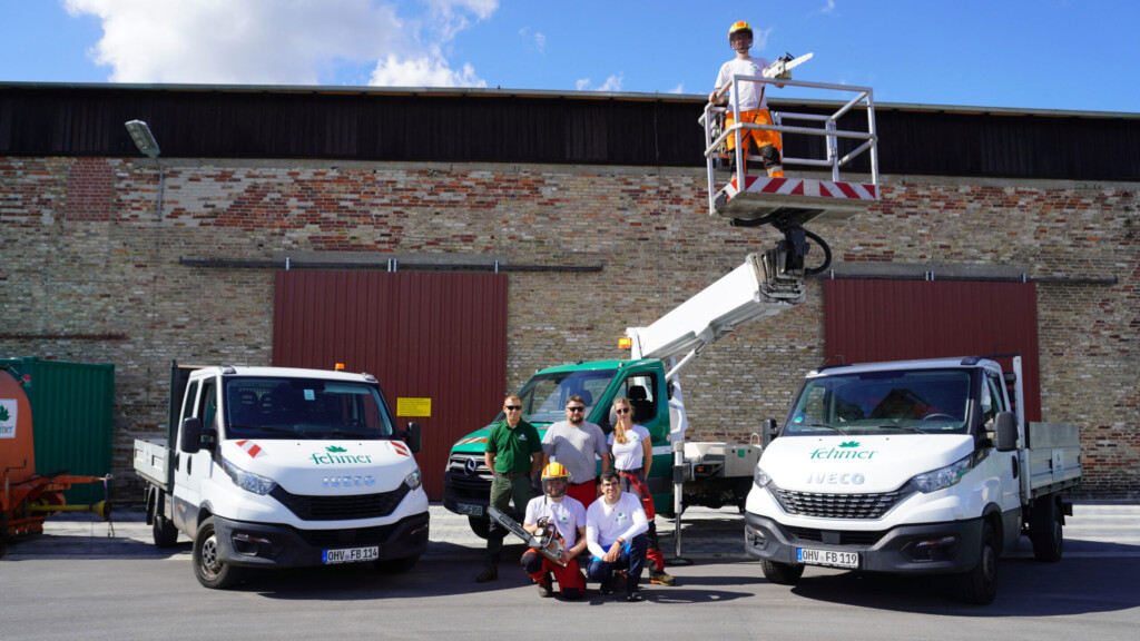 Fünf Personen in Arbeitskleidung vor zwei weißen Fahrzeugen mit dem Logo 'Fehmer', eine Person steht auf einer Hebebühne, im Hintergrund eine Backsteinmauer mit zwei großen Toren.