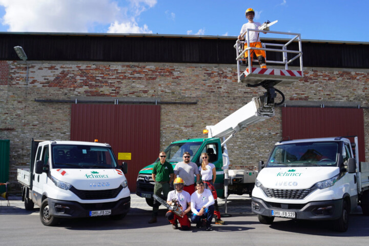 Fünf Personen in Arbeitskleidung vor zwei weißen Fahrzeugen mit dem Logo 'Fehmer', eine Person steht auf einer Hebebühne, im Hintergrund eine Backsteinmauer mit zwei großen Toren.