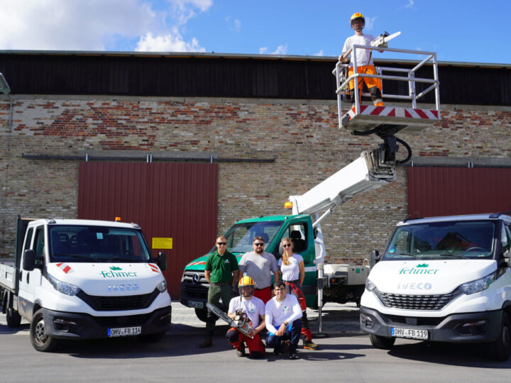 Fünf Personen in Arbeitskleidung vor zwei weißen Fahrzeugen mit dem Logo 'Fehmer', eine Person steht auf einer Hebebühne, im Hintergrund eine Backsteinmauer mit zwei großen Toren.