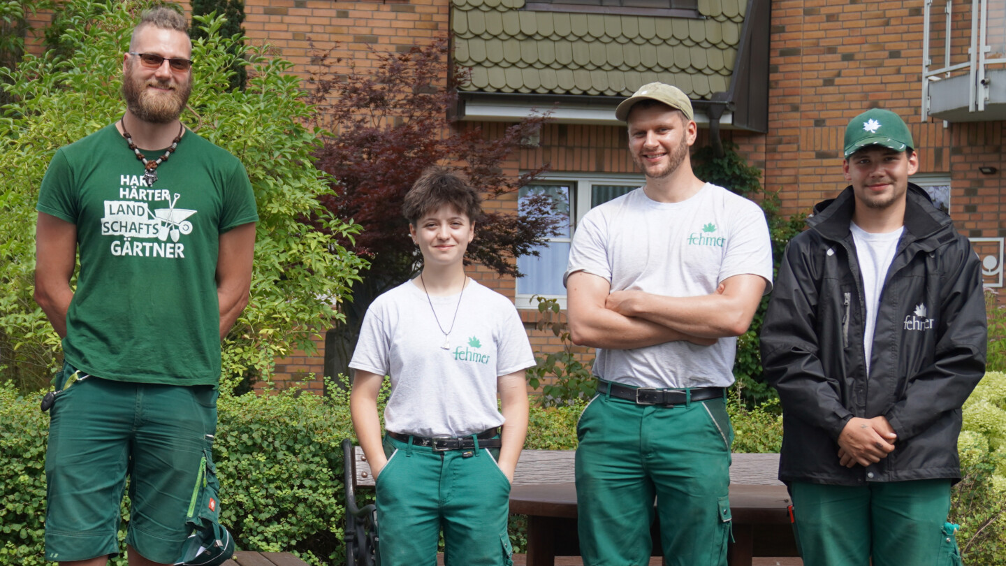 Vier junge Personen stehen nebeneinander vor einem Haus mit Backsteinfassade, drei tragen grüne Arbeitshosen und T-Shirts, einer eine schwarze Jacke und Mütze.