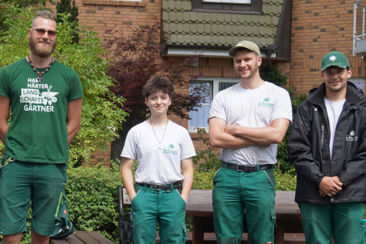 Vier junge Personen stehen nebeneinander vor einem Haus mit Backsteinfassade, drei tragen grüne Arbeitshosen und T-Shirts, einer eine schwarze Jacke und Mütze.