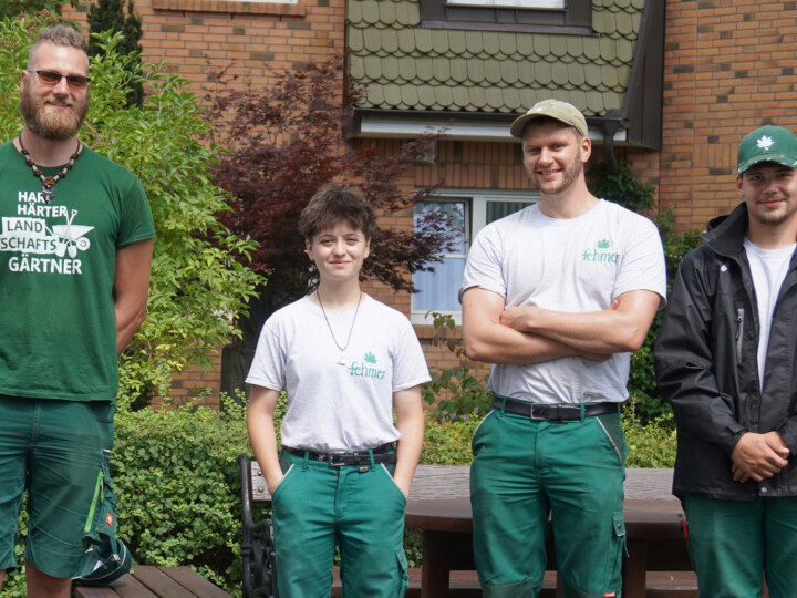 Vier junge Personen stehen nebeneinander vor einem Haus mit Backsteinfassade, drei tragen grüne Arbeitshosen und T-Shirts, einer eine schwarze Jacke und Mütze.