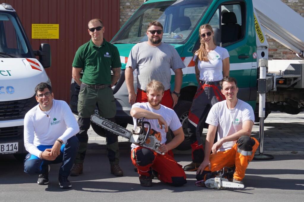 Sechs Personen in Arbeitskleidung posieren vor zwei Fahrzeugen, eine Person hält eine Motorsäge